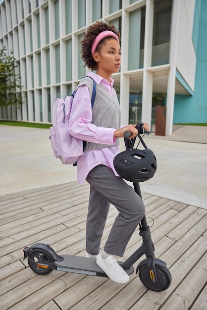 Full length shot of curly haired teenage girl dressed in streetwear carries backpack rides electric scooter at street during free time at weekend