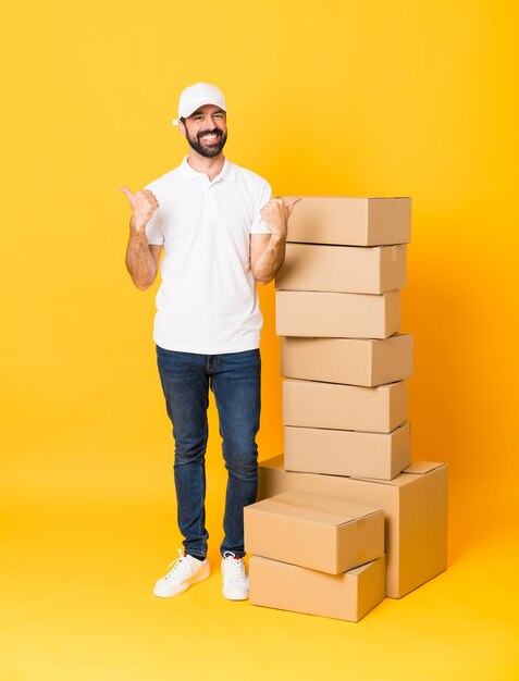 Full-length shot of delivery man among boxes over isolated yellow wall with thumbs up gesture and smiling