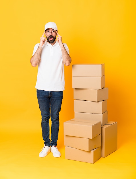 Full-length shot of delivery man among boxes over isolated yellow  with glasses and surprised