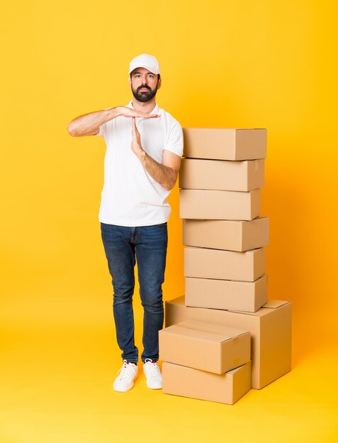 Full-length shot of delivery man among boxes over isolated yellow making time out gesture