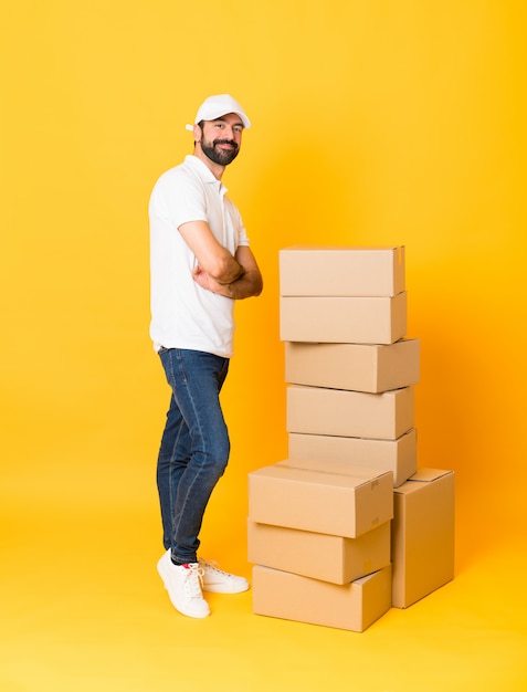 Full-length shot of delivery man among boxes over isolated yellow with arms crossed and looking forward