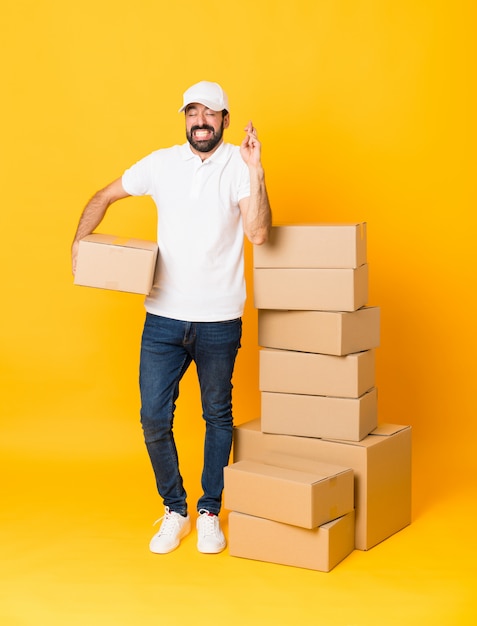 Full-length shot of delivery man among boxes over isolated yellow with fingers crossing and wishing the best