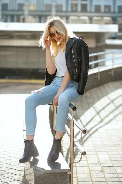 Full length shot of glamor blonde woman wearing vintage glasses sitting on street parapet