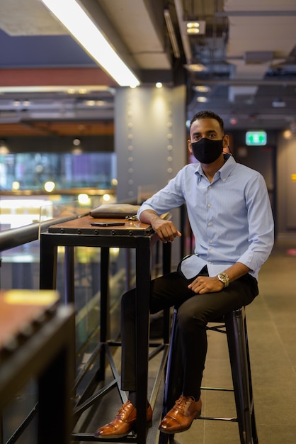 Full length shot of of handsome black African businessman sitting inside shopping mall while wearing face mask to protect from covid-19 vertical shot