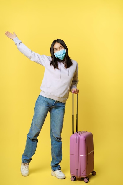 Снимок в полный рост счастливой корейской туристки на отдыхе, позирующей с чемоданом в медицинской маске ...