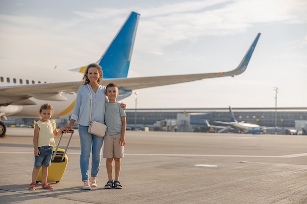 昼間に大きな飛行機の前に立っている間、カメラに微笑んでいる2人の小さな子供と幸せな母親のフルレングスのショット。家族、休暇、旅行のコンセプト