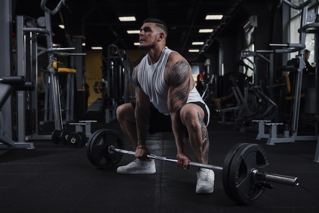 Полнометражный снимок огромного мускулистого татуированного фитнесмена, тренирующегося со штангой в спортзале
