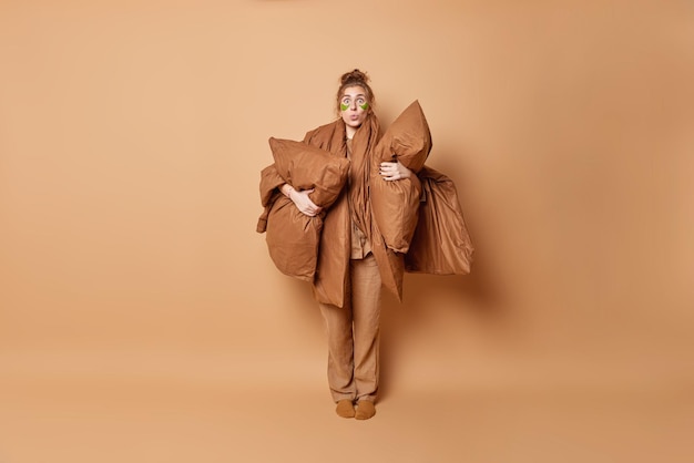 Full length shot of impressed woman with messy hair gathered in
bun wears pajama holds blanket and pillows applies green patches
under eyes prepares for sleep isolated over beige background.