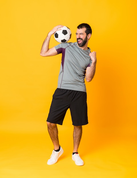 Full-length shot of man over isolated yellow background with soccer ball celebrating a victory