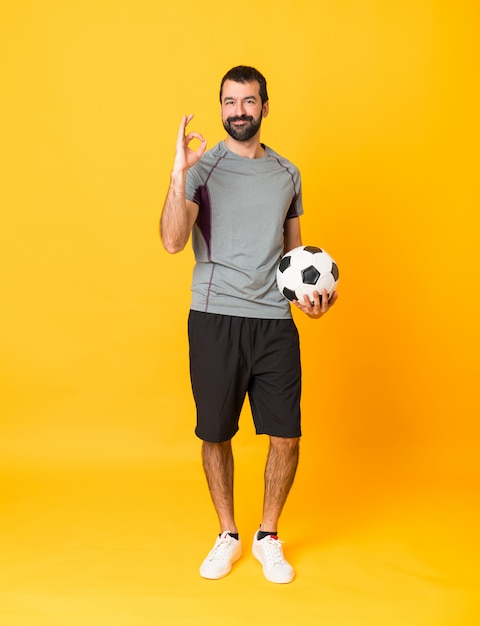 Full-length shot of man over isolated yellow background with soccer ball and making OK sign