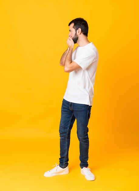 Full-length shot of man with beard over isolated yellow background covering mouth and looking to the side