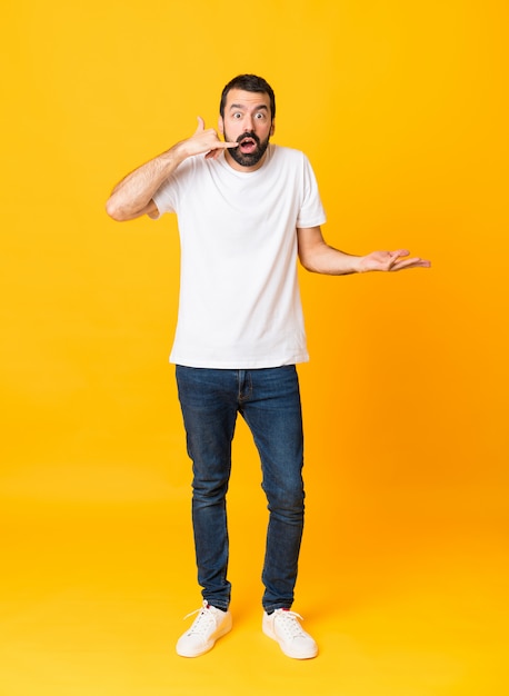 Full-length shot of man with beard over yellow making phone gesture and doubting