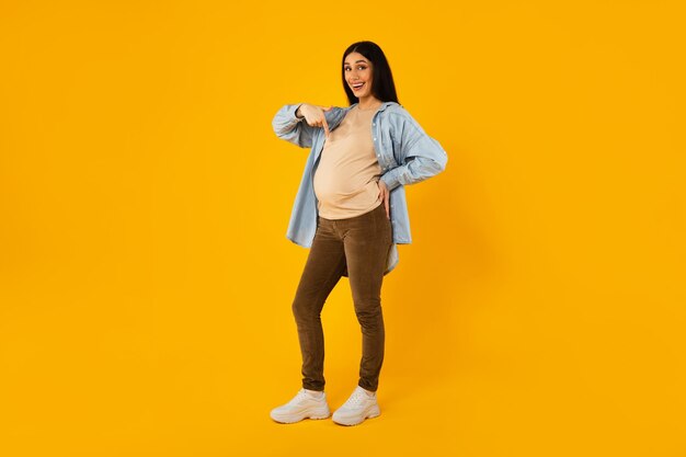 Full length shot of pregnant woman pointing at her big belly showing tummy and smiling yellow background free space