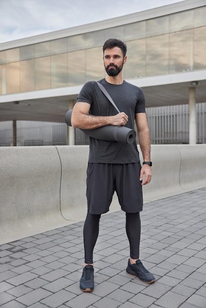 운동할 준비가 된 피트니스 매트가 있는 스포츠맨 포즈의 전체 길이 샷은 흐릿한 도시 배경에 대해 거리 스탠드로 결정된 근육질 팔을 가지고 있습니다. 운동 남자는 야외에서 운동