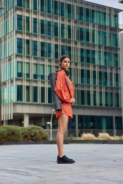 Full length shot of sporty muscular woman dressed in anorak and\
shorts carries rolled karemat for fitness mat looks seriously at\
camera poses against urban building healthy lifestyle concept