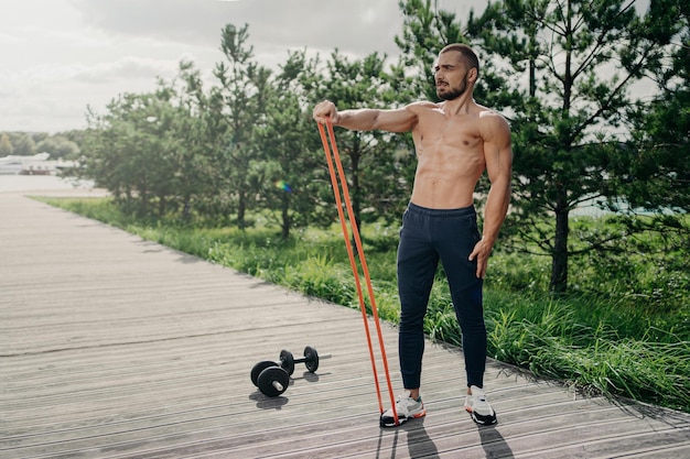 Full length shot of strong bearded sportsman flexes muscles with elastic resistance band wears sport trousers and sneakers stands outdoor uses sport equipment for workout performs exercises