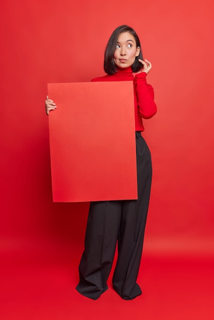 Full length shot of thoughtful Asian woman holds banner for your advertising content or promotional text wears turtleneck and black formal trousers poses against red wall