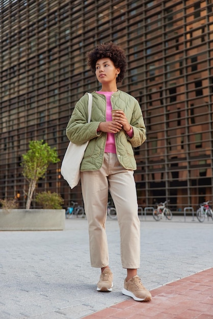 Photo full length shot of thoughtful curly haired young woman holds paper cup of aromatic coffee looks away pensivelyy dressed in street outfit waits for someone poses outdoors against blurred background