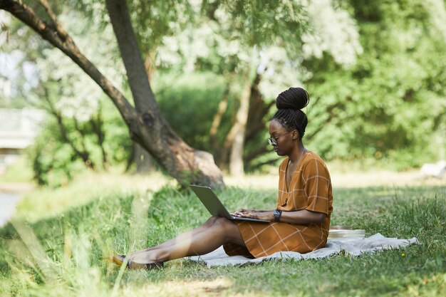 水辺に座って公園でラップトップを使用して、アフリカ系アメリカ人の若い女性の完全な長さの側面図、コピースペース