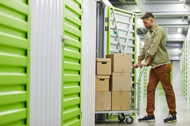 Photo full length side view at handsome bearded man loading cart with cardboard boxes into self storage unit, copy space