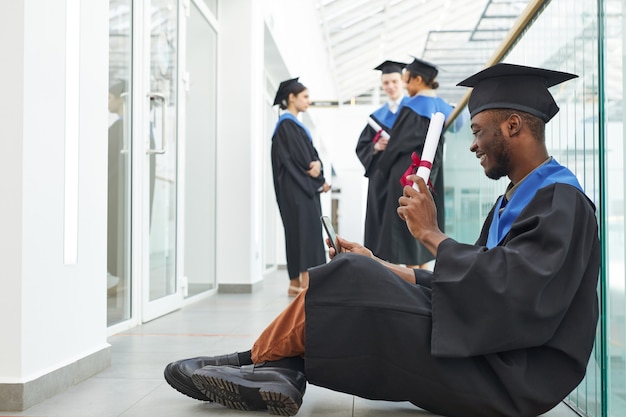 졸업 가운을 입고 바닥에 앉아 영상 채팅을 할 수 있는 졸업장을 보여주는 행복한 아프리카계 미국인 남성의 전체 길이 측면 보기