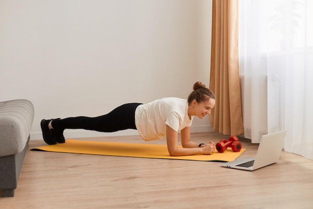 Full length side view portrait of athletic woman wearing white T-shirt and black leggins training her abdominal muscles, standing in plank pose, fitness at home on yoga mat.