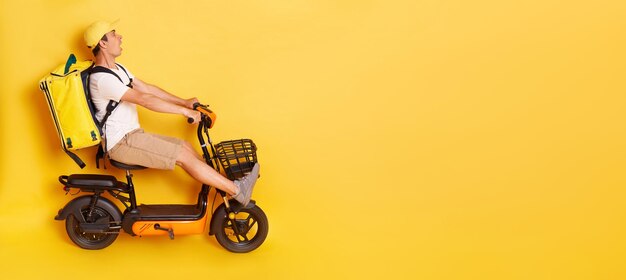 Full length side view portrait of man courier with thermo backpack in white Tshirt and cap riding electric bike braking avoids accident looks scared and shocked isolated over yellow background