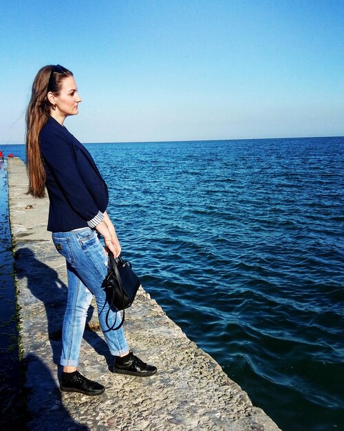 Full length side view of woman standing by blue sea against clear sky