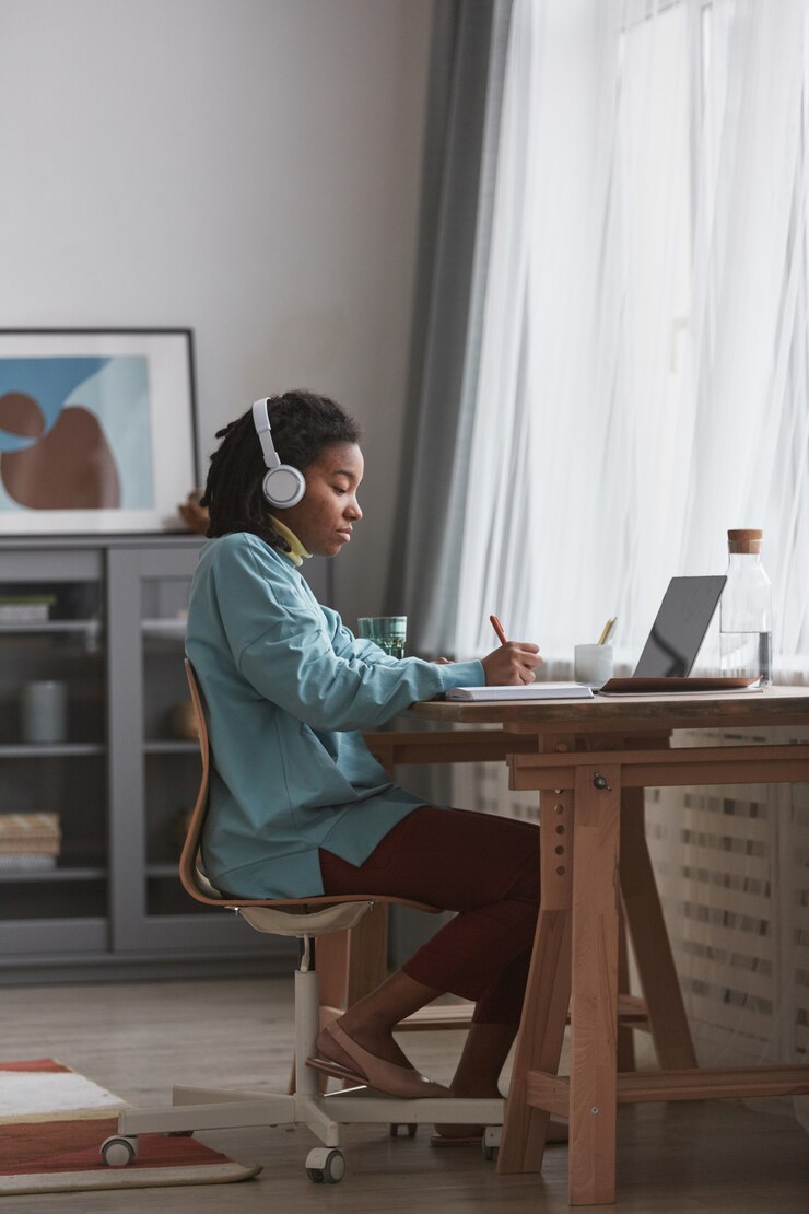 Photo full length side view at young african american woman studying at home or working at desk while wearing headphones