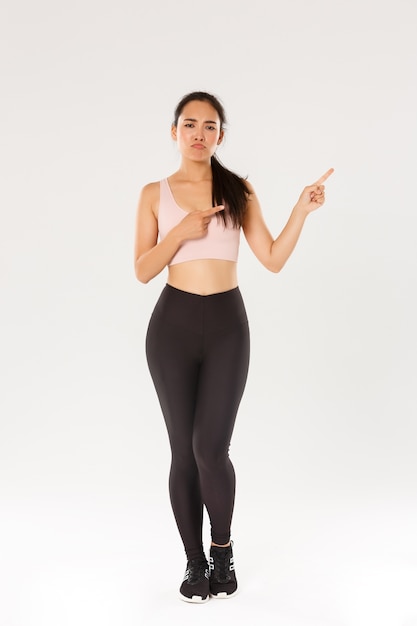 Full length of skeptical, disappointed asian fitness girl, sportswoman smirk and sulking while complaining on bad gym or awful workout gear, pointing upper right corner displeased, white background.