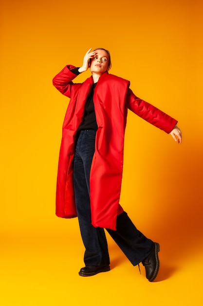 Full length slim woman in trendy oversize coat looking away and dancing against yellow background