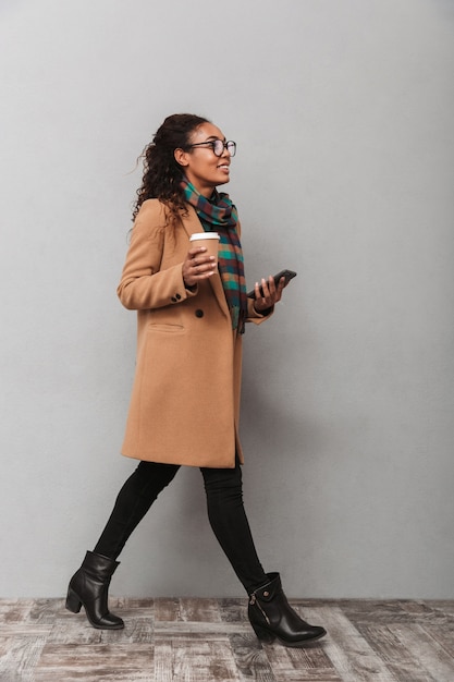 Full length of a smiling african woman wearing coat walking, holding mobile phone, drinking takeaway coffee