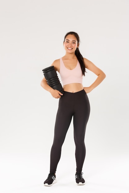 Full length of smiling good-looking asian fitness coach in sportswear looking pleased, holding foam roller, reducing soreness after productive training session, roll through tight muscles.