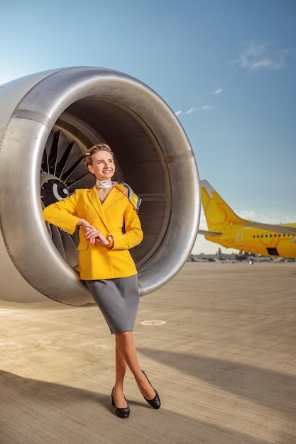 Full length of smiling woman flight attendant wearing yellow jacket while standing near aircraft turbine engine