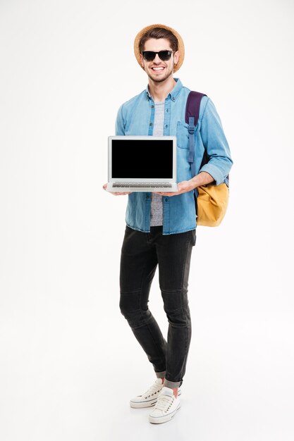 空白の画面のラップトップを立って保持しているバックパックと笑顔の若い男の全長