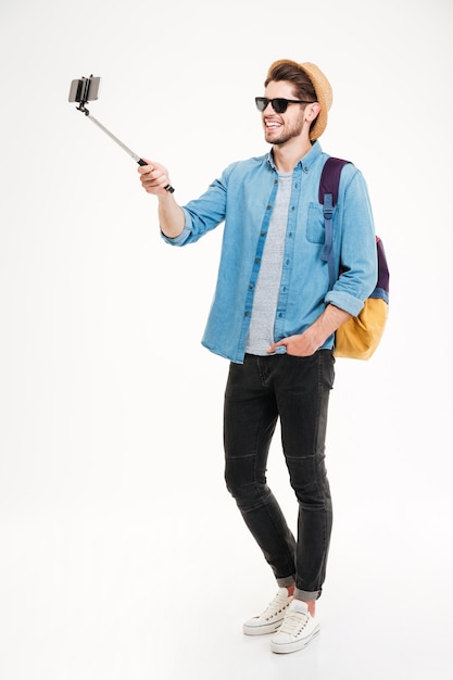 Full length of smiling young man with backpack taking photos with smartphone and selfie stick