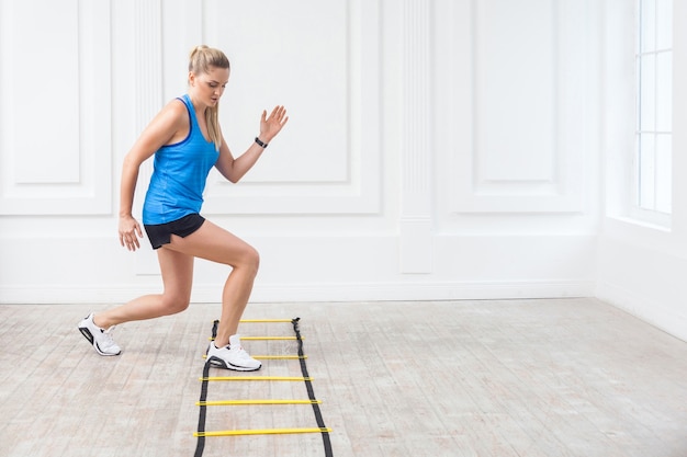 Full length of sporty beautiful young athletic blonde woman in black shorts and blue top are hard working and training on agility ladder drills in gym. Indoor, studio shot, workout and sport concept.