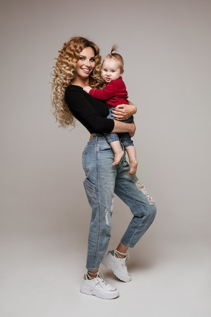 Full length stock photo of gorgeous blonde Caucasian mother in jumper and ripped jeans and sneakers holding her baby daughter in arms, smiling at camera.