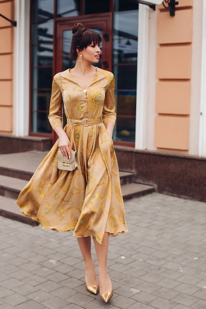 Photo full length stock photo of a gorgeous elegant lady in golden dress, high heels of golden color and crossbag. she is walking in the street smiling.