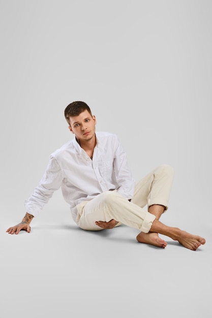 Full length studio portrait of young barefoot man in white shirt and ivory trousers sitting the floor