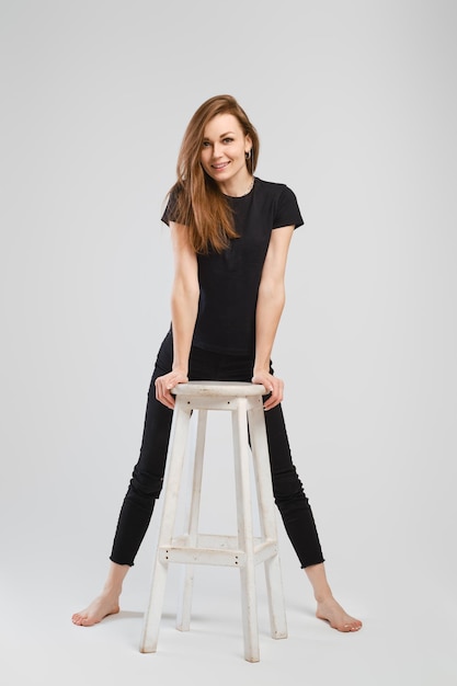 黒のジーンズと背の高い椅子に寄りかかってシャツを着た若い裸足の女性の完全な長さのスタジオの肖像画