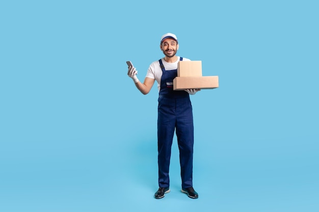 Full length surprised delivery man in uniform and gloves holding mobile phone cardboard boxes and looking at camera with amazed expression Courier shocked by online order studio shot isolated