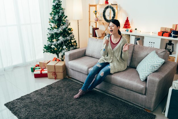 full length taiwanese girl is spending Christmas holiday singing koraoke. woman sitting in the decorated room with microphone is having party by herself. authentic lifestyle