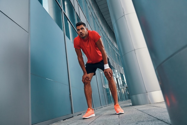 Photo full length of tired young man in sports clothing making a break during his sport training while exercising outdoors