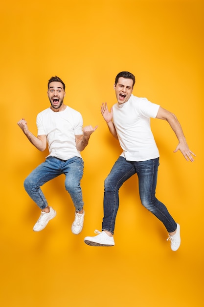 Full length of two cheerful excited men friends wearing blank t-shirts jumping isolated over yellow wall, celebrating