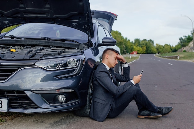 Full length of unhappy young businessman using cell phone by broken down car at countryside