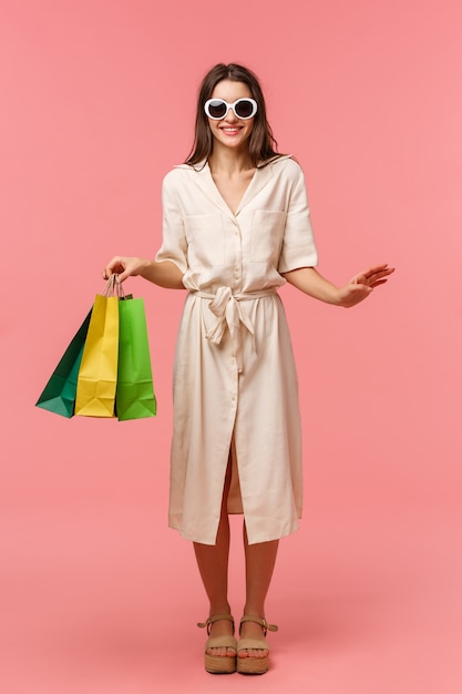 Full-length verticaal portret elegante knappe vrouw op vakantie, winkelen en plezier maken in winkelcentra, zonnebril dragen, lichte jurk, draagtassen uit winkel, roze muur staan glimlachen