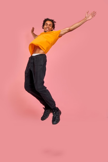 Вертикальное фото в полный рост стильного счастливого темнокожего мужчины празднует поднятые вверх руки, широко прыгая в воздухе, на розовом фоне. Выразительная спешка, распродажа, покупки. Эмоции, рекламная концепция