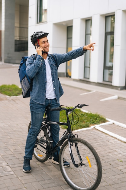 Full length vertical portrait of cheerful handsome bicycle delivery man with thermal backpack talking on mobile phone, sees customer and marks location by raising hand.