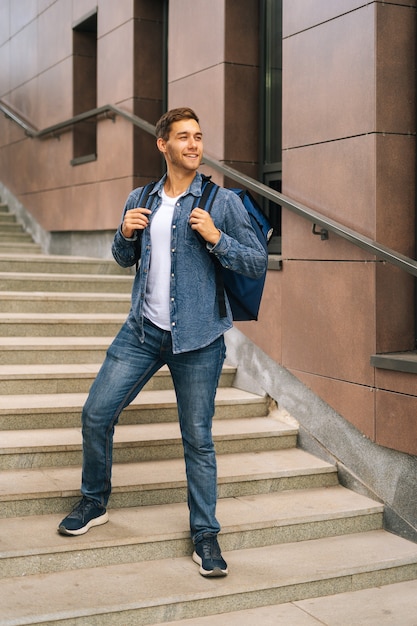 Full length vertical portrait of handsome young male of food delivery service with large thermal backpack standing posing in office building stairs, blurred background, looking away.